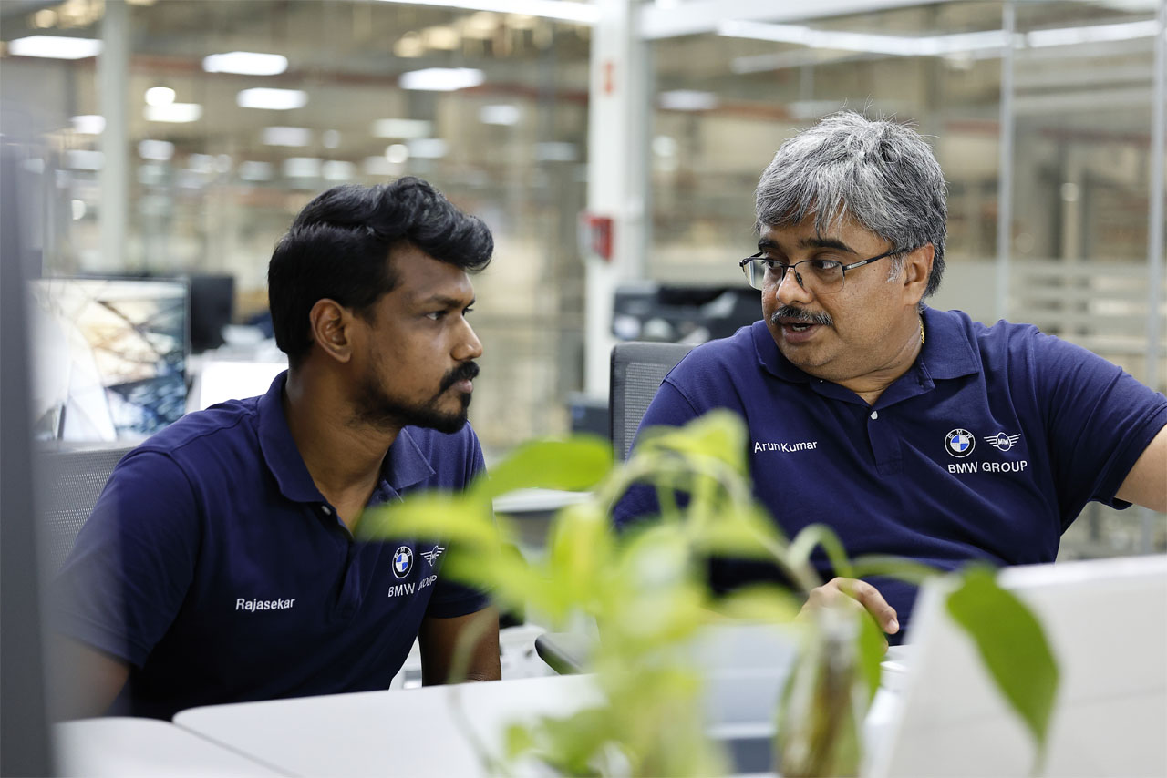 Two men in blue shirts are seated at a table, working on laptops.