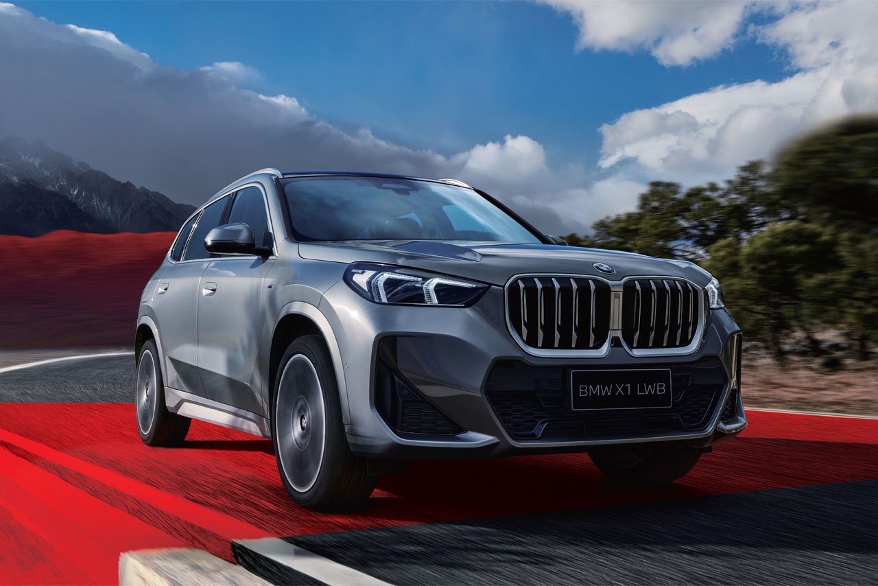 BMW X1 SUV driving on a winding road with red pavement under a cloudy sky and mountains in the background.