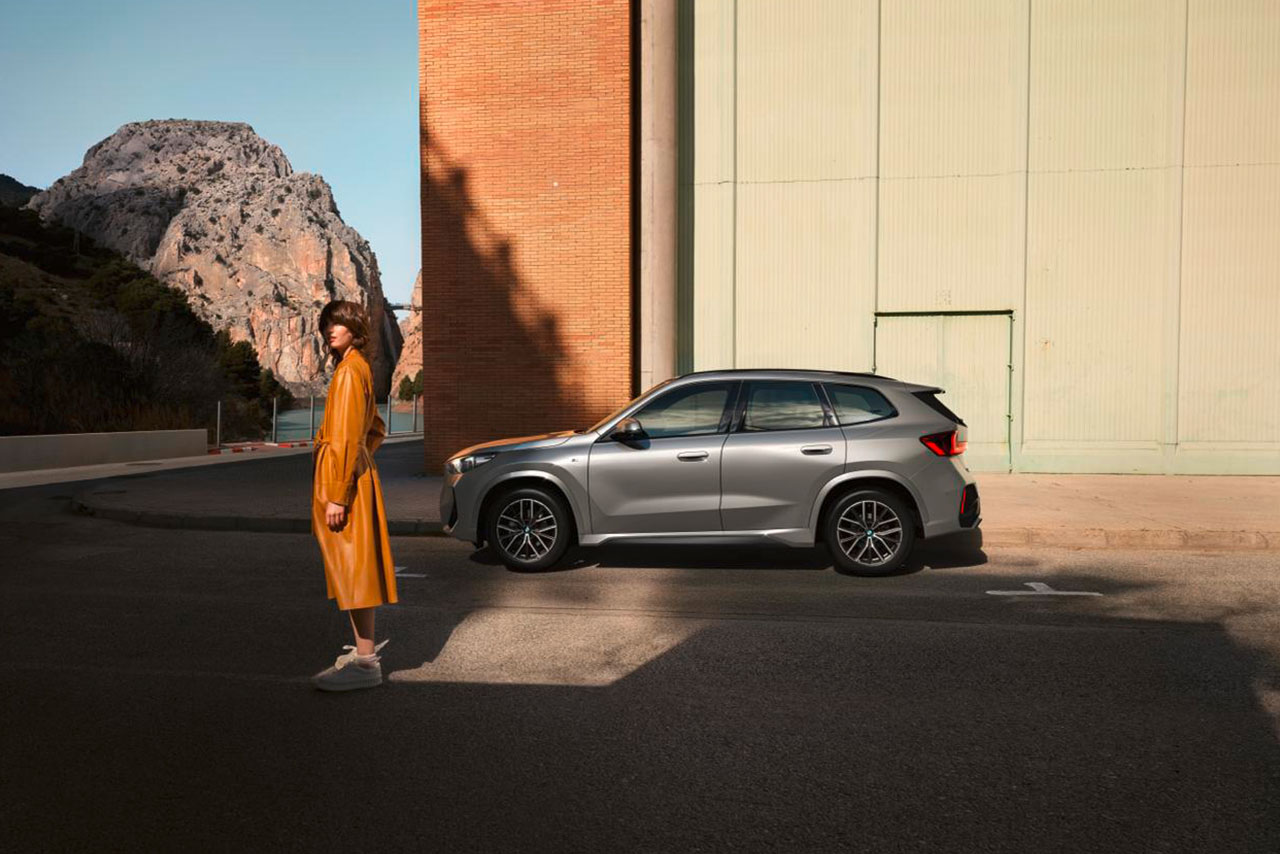 A woman in an orange coat standing infront of an Matte grey BMW X1.