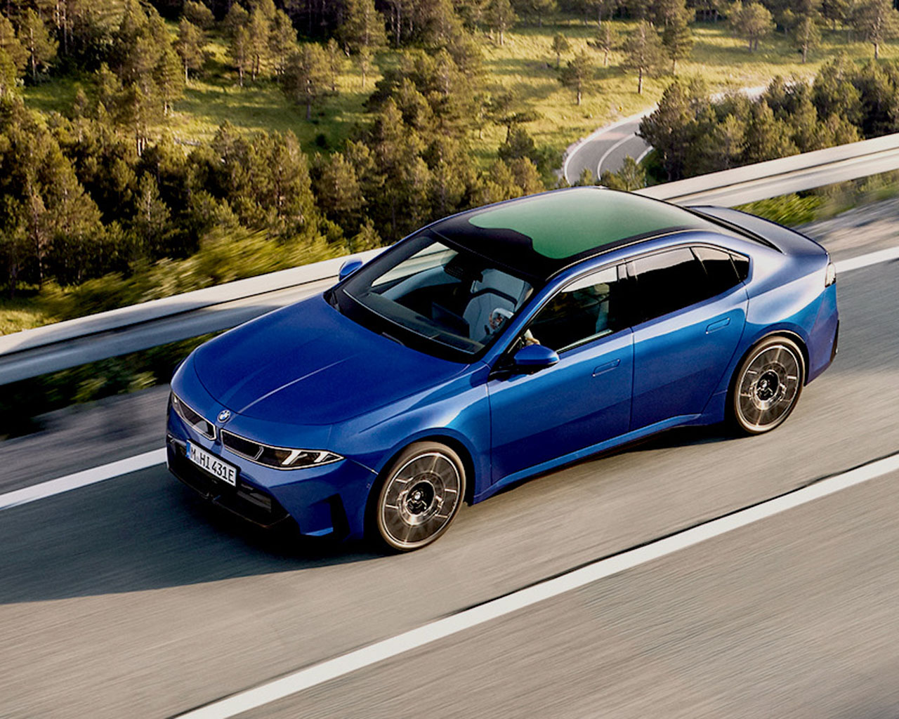  Blue BMW iX3 driving along a winding country road through a forested landscape.
