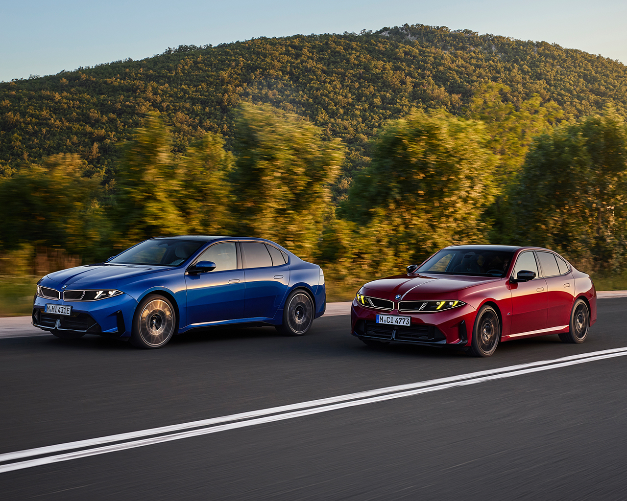 A blue fully electric BMW i3 50 xDrive (left) and a red BMW M350 xDrive drive dynamically side by side, slightly staggered, on a country road, viewed from a front three-quarter perspective, with a forested mountain landscape in the background.