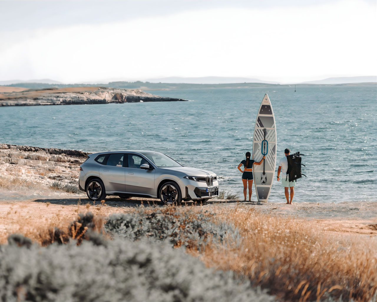 A man and woman with a surfboard next to a BMW iX3 by the sea.