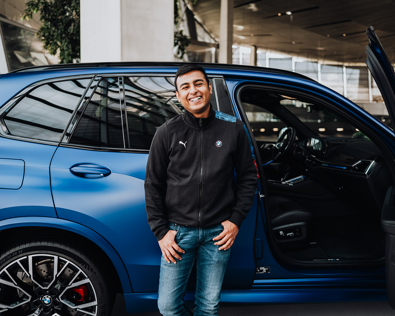 Trainee Kartikey lachend vor einem blauen BMW X5M