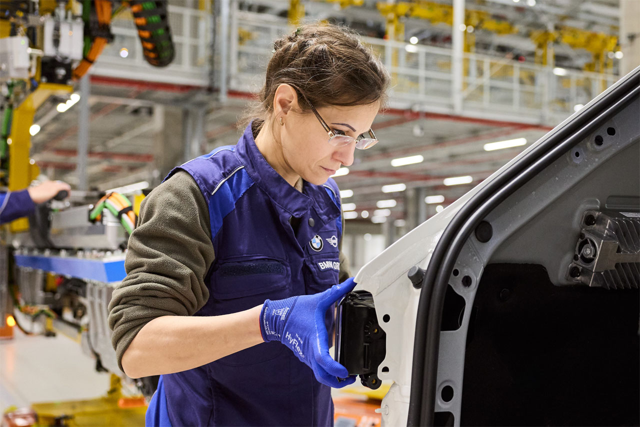 A woman with workclothes is fixing parts to a car body in production.