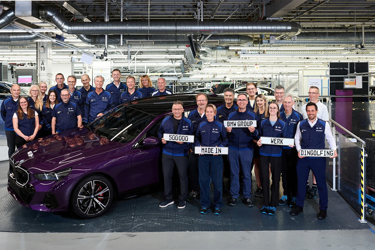 Gruppe von Personen in blauer Arbeitskleidung steht neben einem dunkelvioletten Auto in einer Fabrikhalle, einige halten Schilder mit den Worten '500000', 'BWM GROUP', 'MADE IN', 'WERK', 'DINGOLFING'.