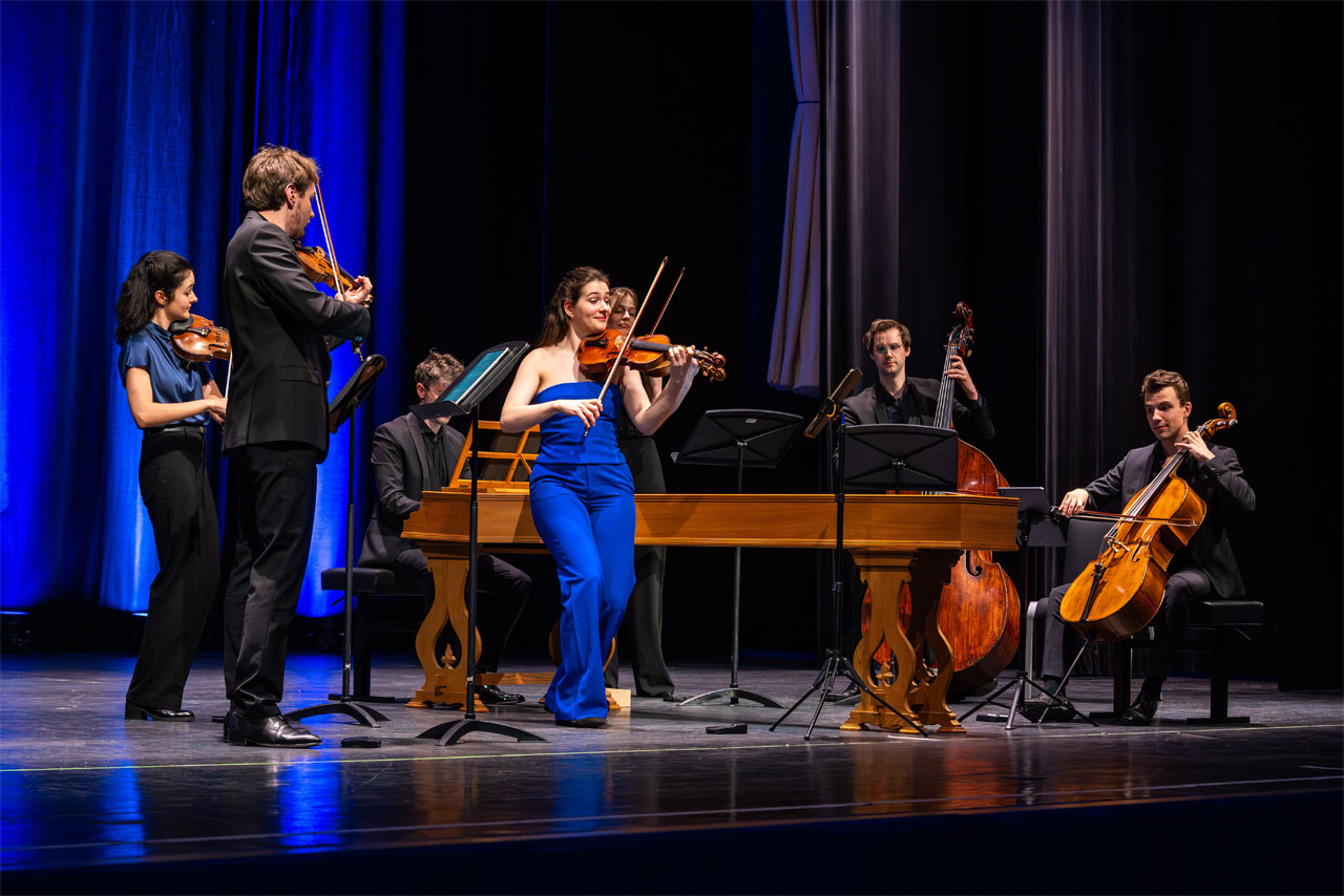 Gruppe von Musikern auf einer Bühne mit Streichinstrumenten und einem Cembalo im Hintergrund.