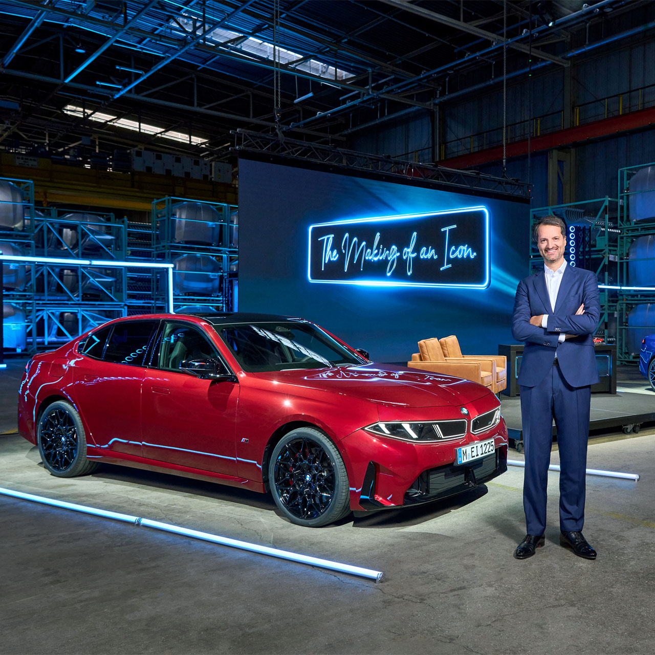 Roter BMW in einer Halle vor einer beleuchteten Tafel mit der Aufschrift 'The Making of an Icon' neben einem Mann im Anzug mit verschränkten Armen.