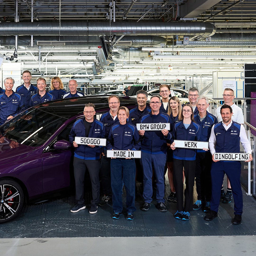Gruppe von Personen in blauer Arbeitskleidung steht neben einem dunkelvioletten Auto in einer Fabrikhalle, einige halten Schilder mit den Worten '500000', 'BWM GROUP', 'MADE IN', 'WERK', 'DINGOLFING'.