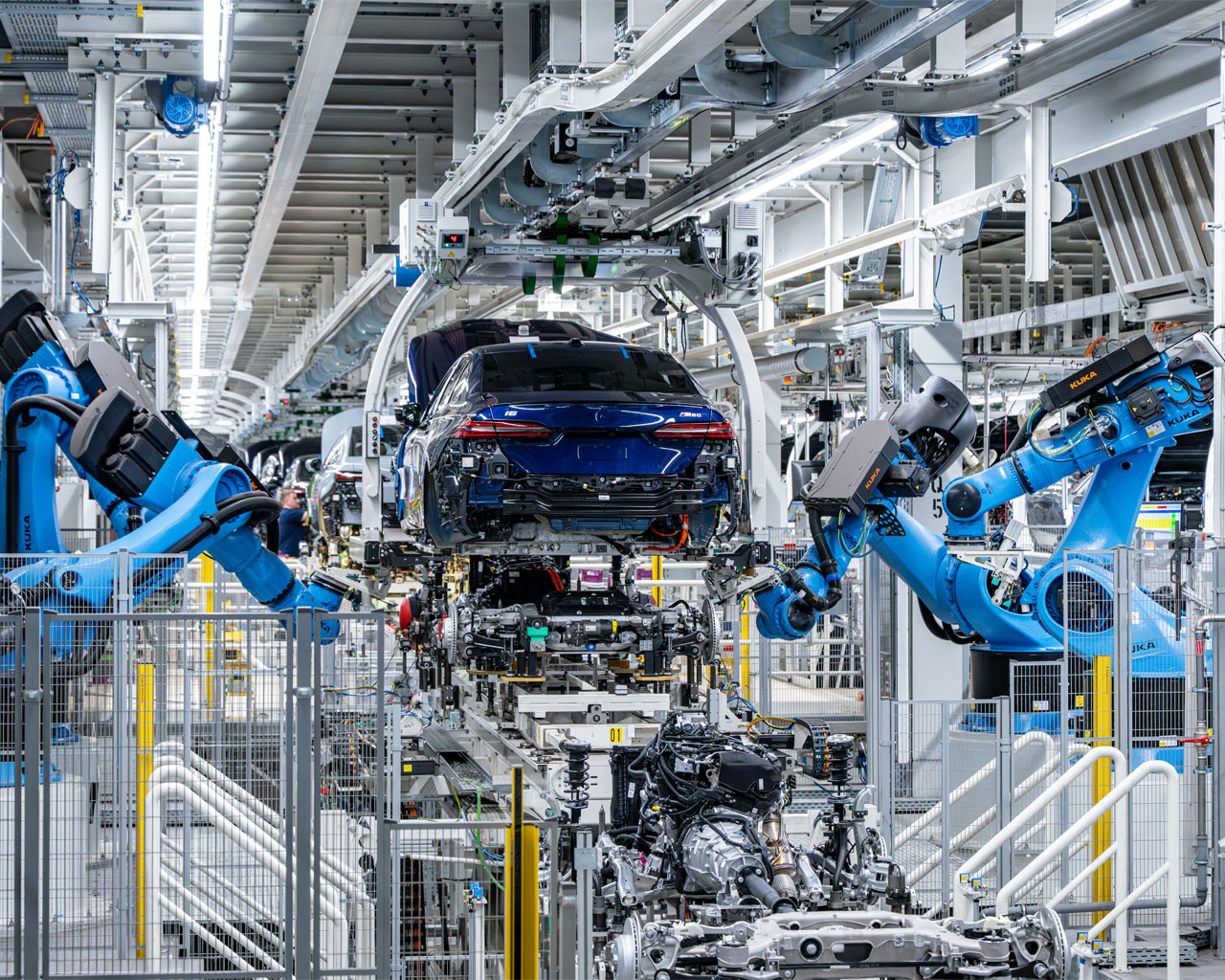 Fabrikhalle mit zwei großen Industrierobotern, die an einem blauen Autoheck arbeiten.