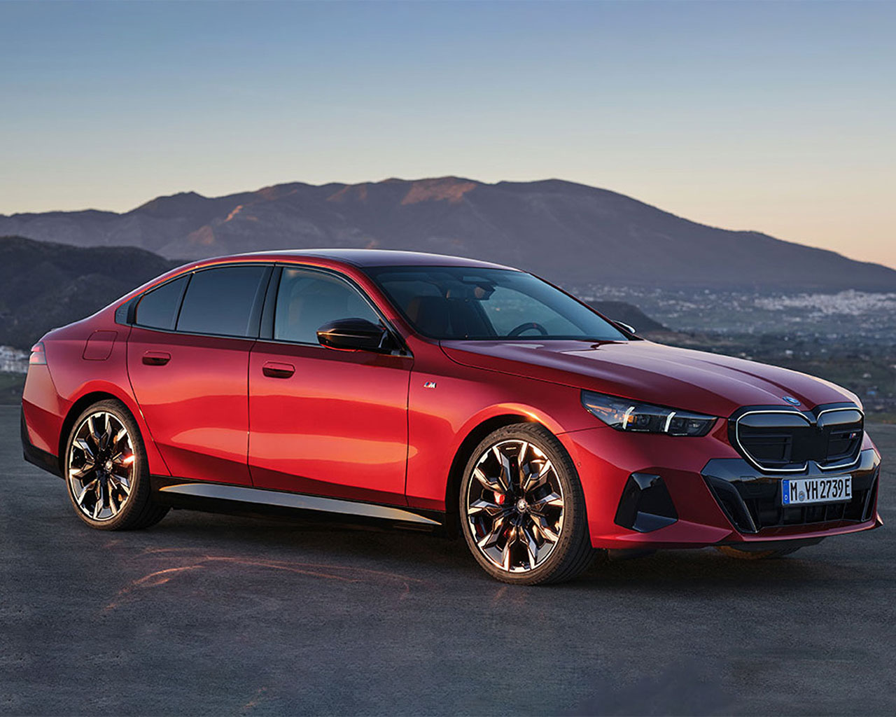Red BMW 5 Series sedan stands elevated above a city at sunset.