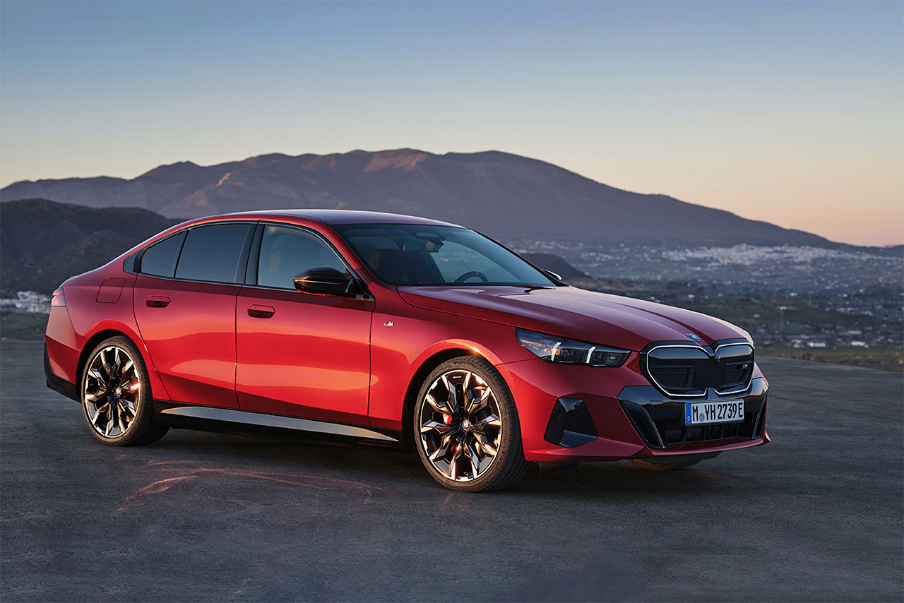 Red BMW 5 Series sedan stands elevated above a city at sunset.