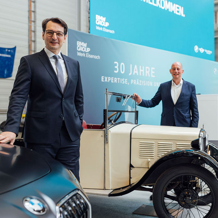 Zwei Männer in Anzügen stehen neben einem historischen BMW-Auto in einer Werkhalle mit blauem Banner '30 Jahre Expertise, Präzision'.