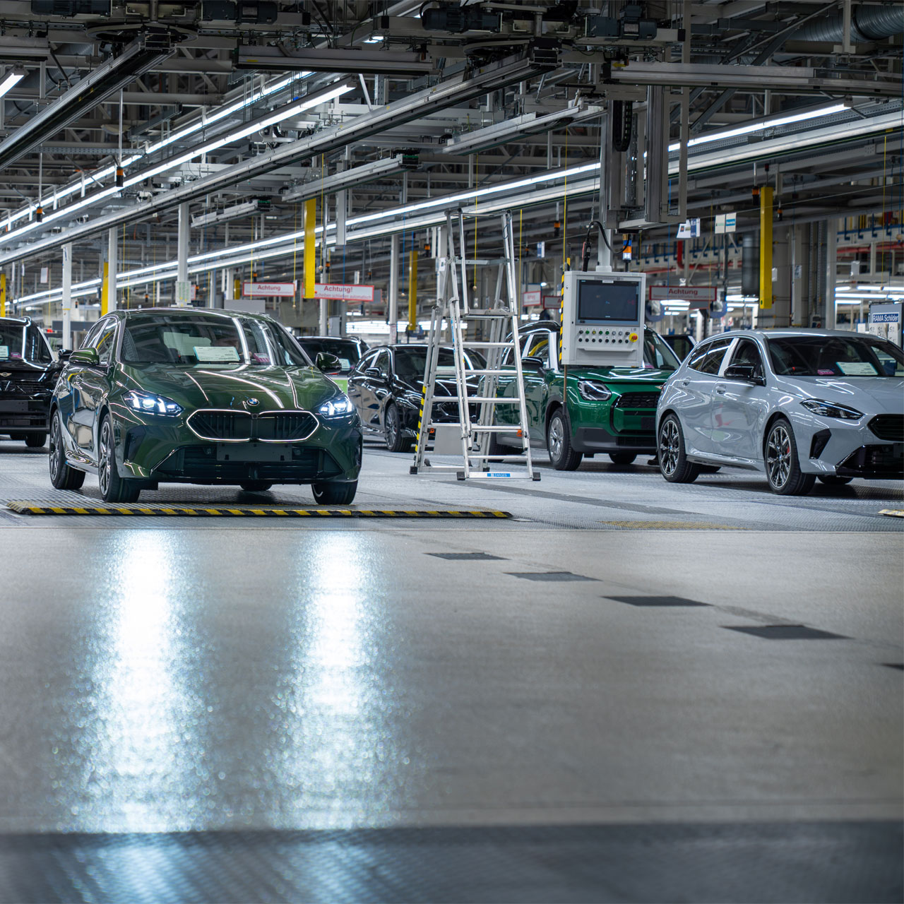 Fabrikhalle mit mehreren in einer Reihe stehenden Autos und technischen Anlagen an der Decke.