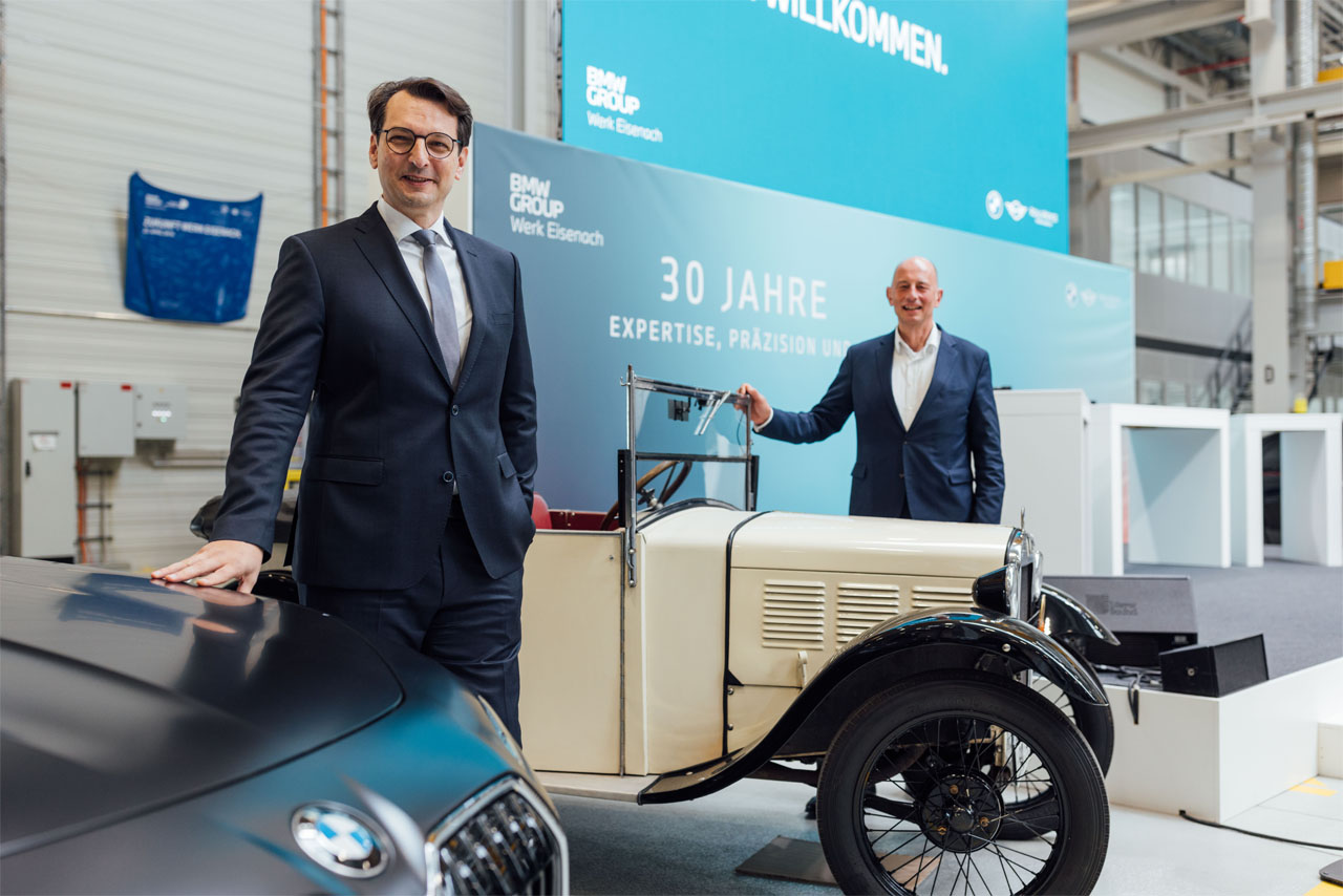 Zwei Männer in Anzügen stehen neben einem historischen BMW-Auto in einer Werkhalle mit blauem Banner '30 Jahre Expertise, Präzision'.