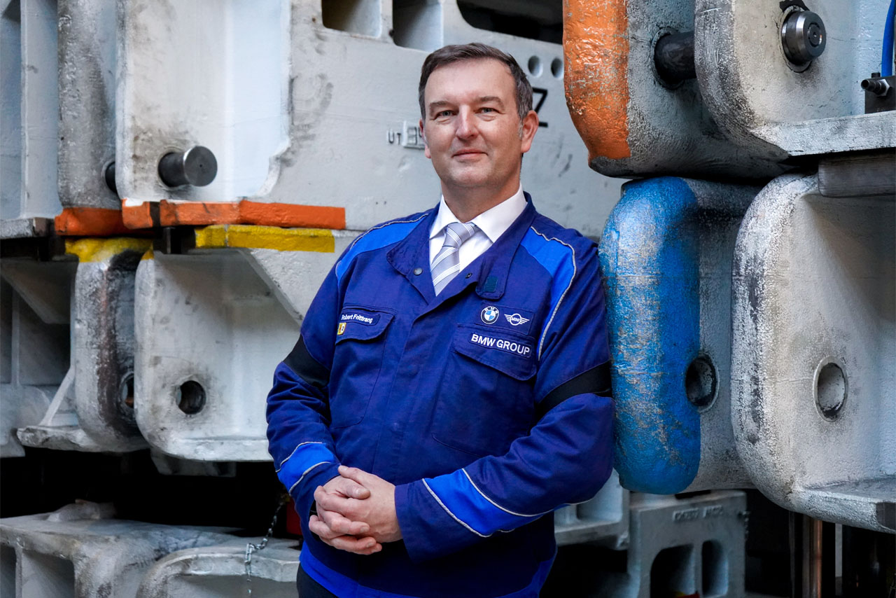 Mann in blauer Arbeitsjacke mit BMW-Group-Logo lehnt an großen, farbigen Metallteilen in einer Werkhalle.