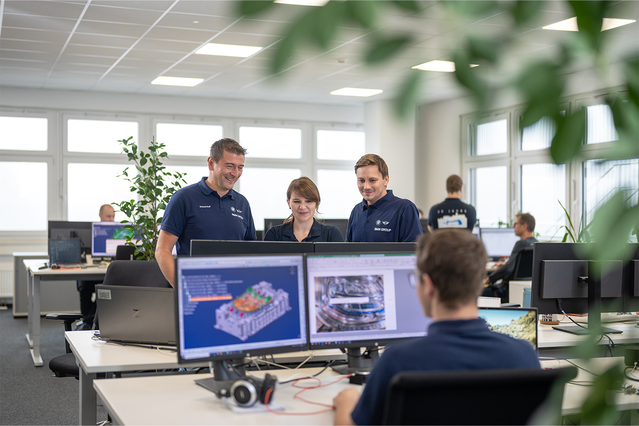 An employee is working at his computer in the office. Three colleagues are standing opposite him, discussing something.