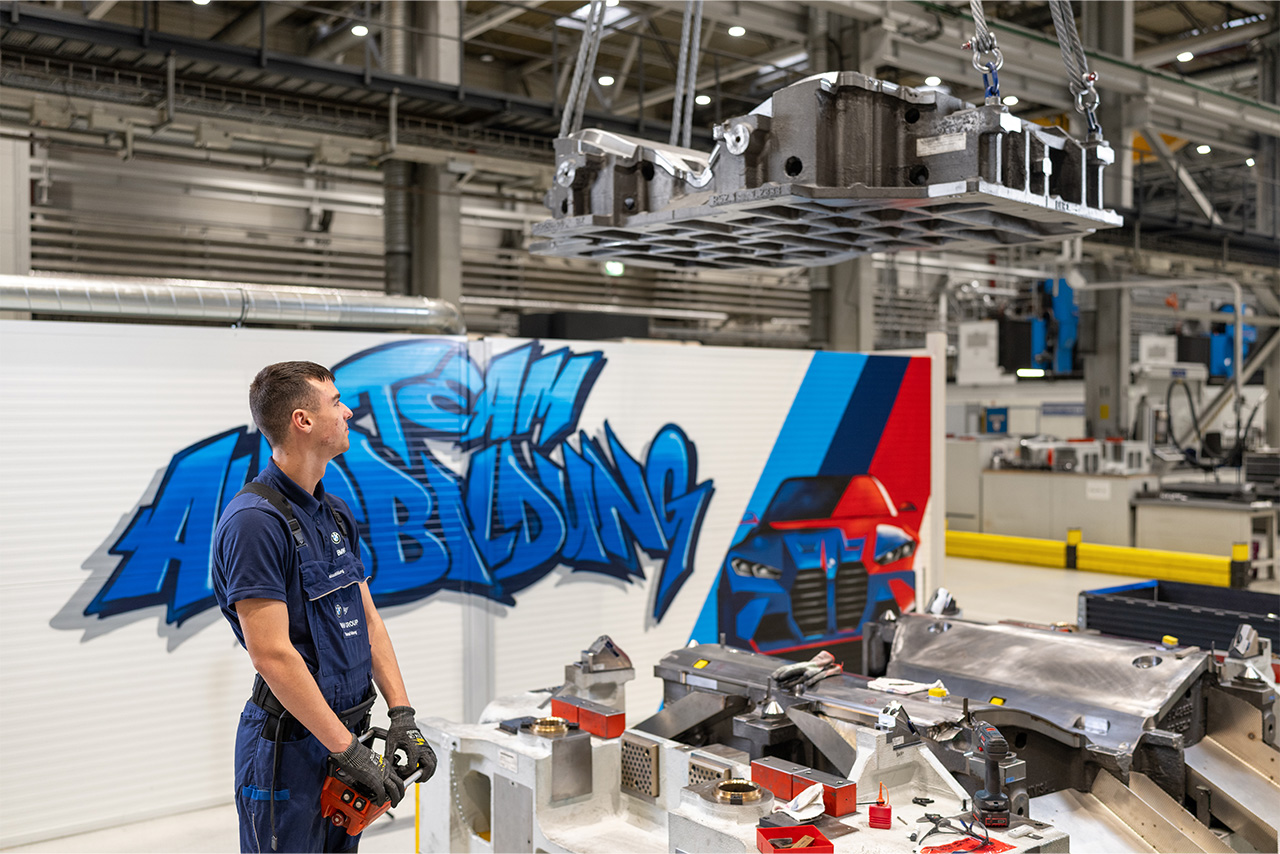 A trainee stands in front of a painted wall with the inscription "Team Ausbildung" and a BMW vehicle on it in production.
