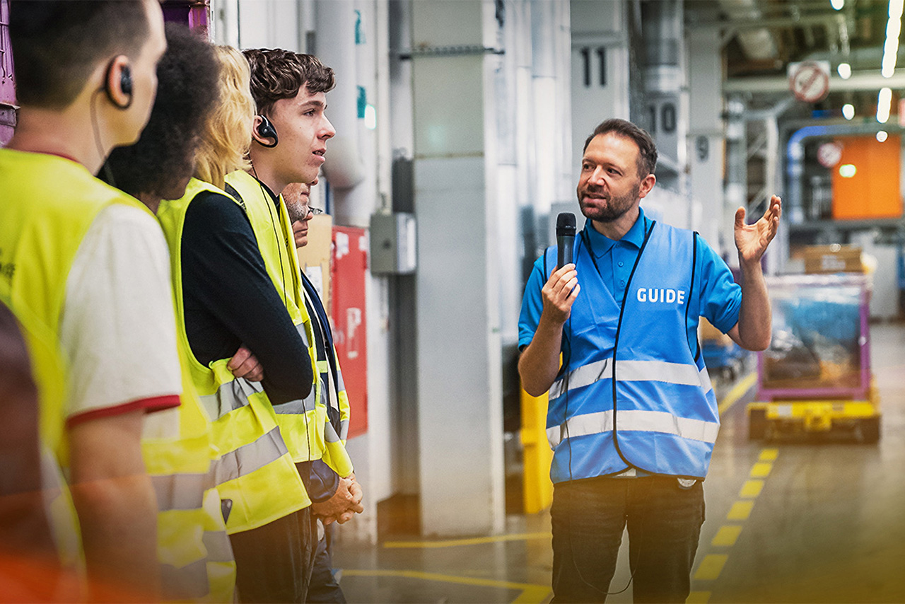 The BMW Guide can be seen from the front. He is holding a microphone in his left hand and pointing at something with his right hand. The visitors are standing to his left.