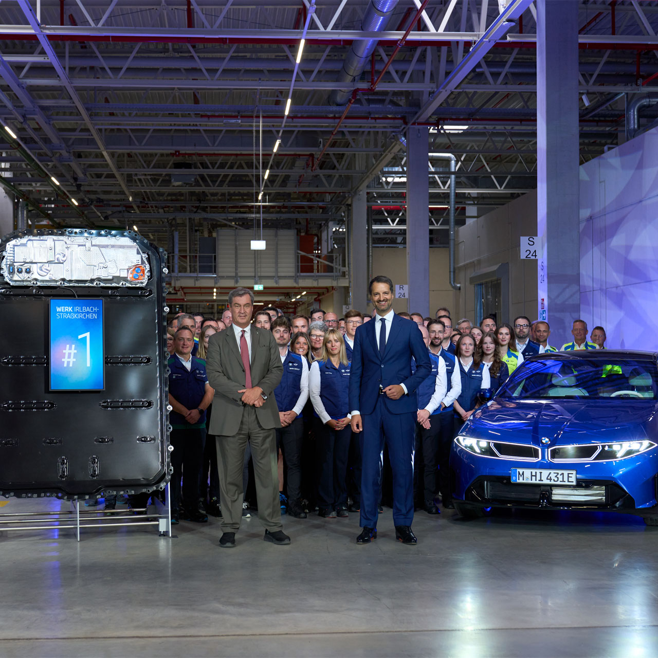 Gruppe von Menschen in Arbeitskleidung und Anzügen steht in einer Fabrikhalle neben einem blauen Auto und einer großen Maschine mit beleuchtetem Display '#1'