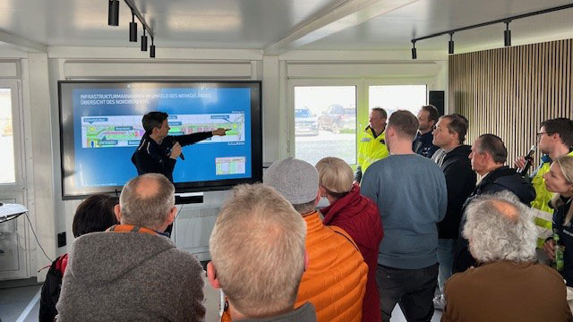 Person zeigt mit Mikrofon auf Bildschirm mit Präsentation vor einer Gruppe Zuhörender in einem Raum.