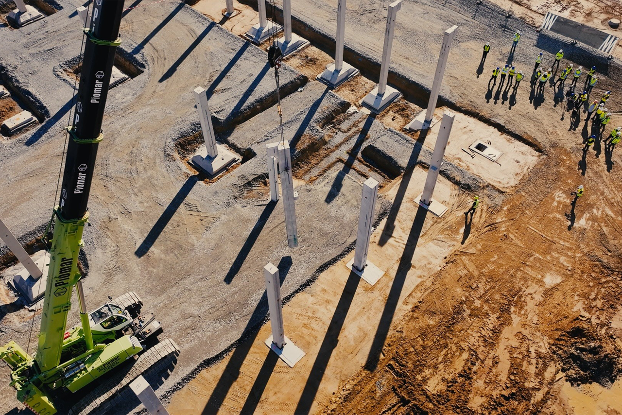 A large crane places support pillars on the construction site while a group of construction workers stand next to it and watch the work.