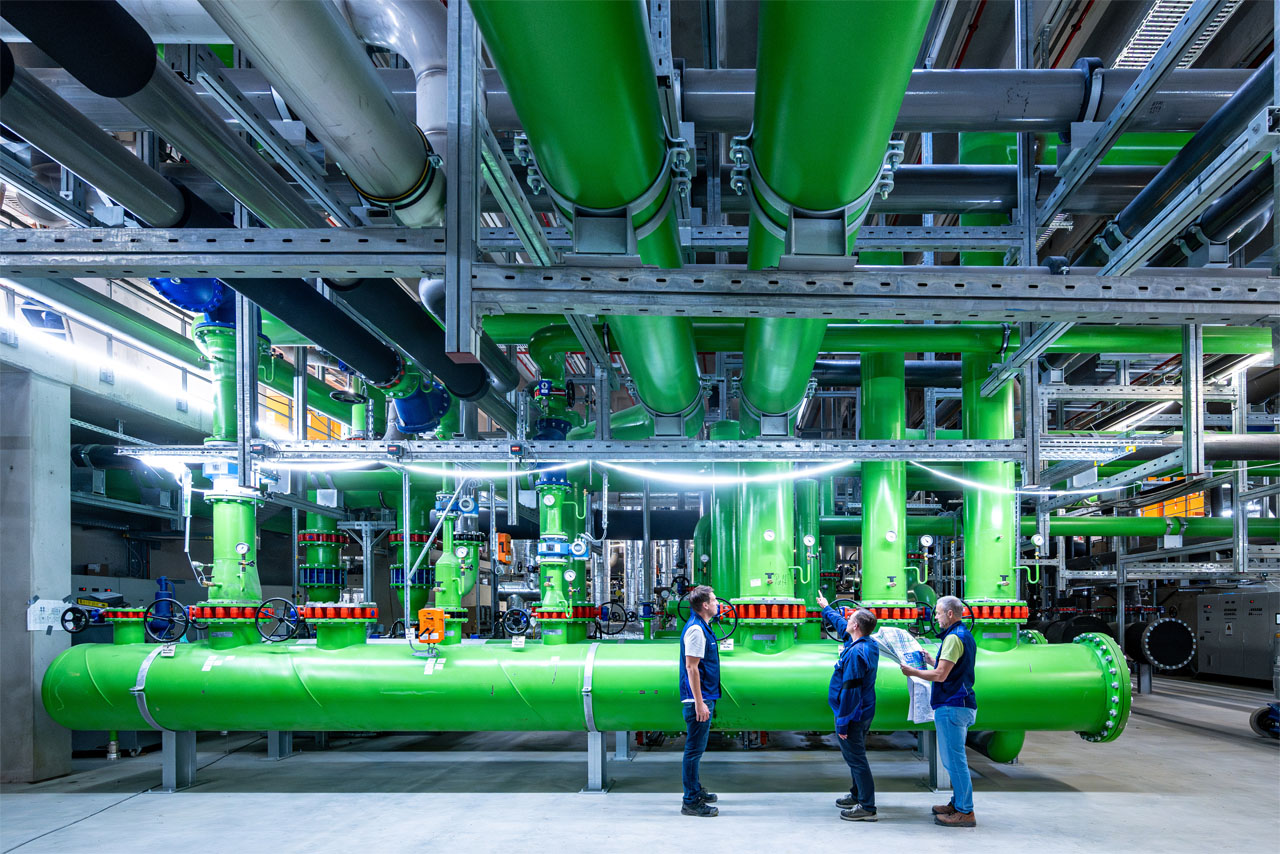 Three people wearing hard hats and safety vests stand in front of large green pipes in an industrial facility.
