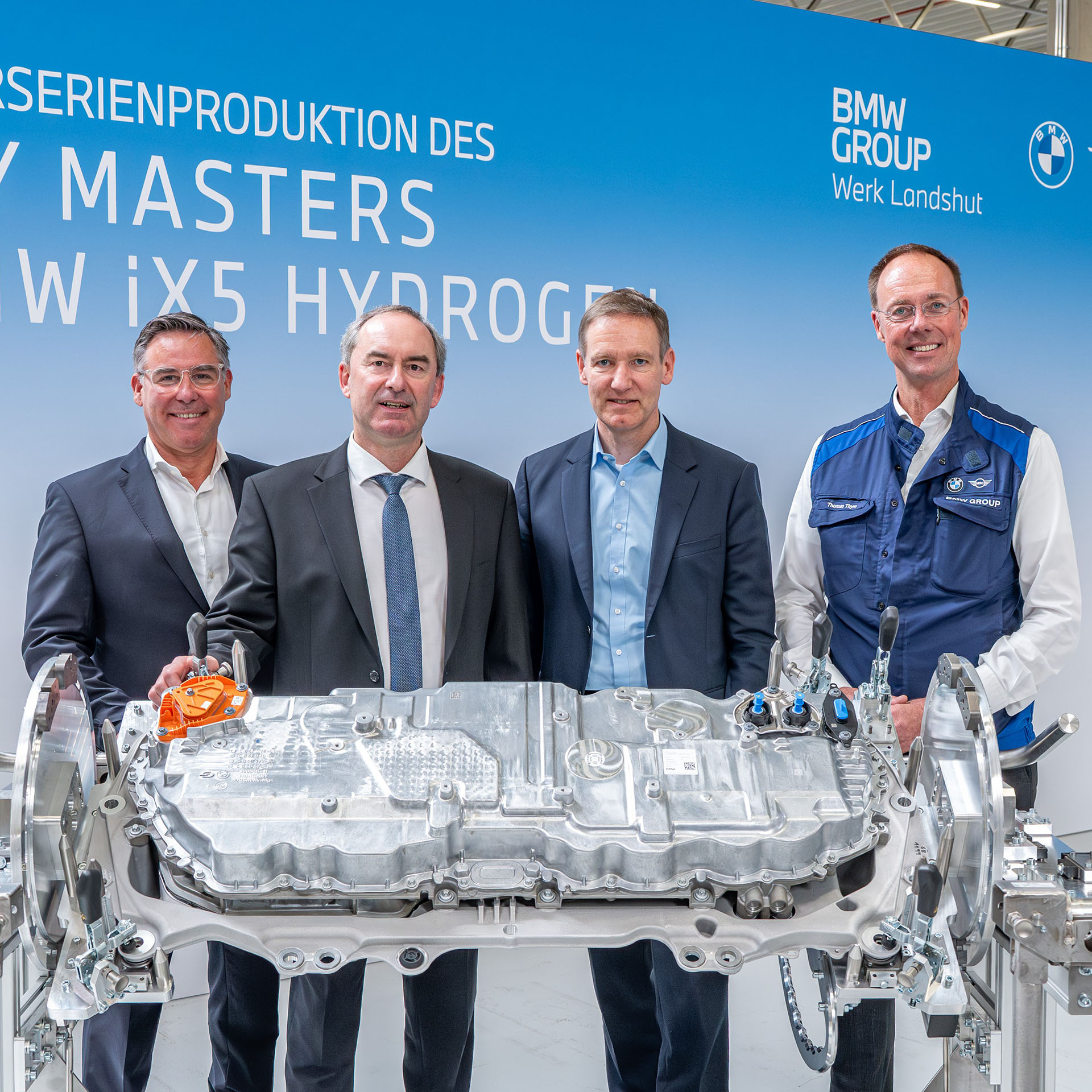Vier Männer in Anzügen und Arbeitskleidung stehen hinter einem großen technischen Bauteil vor einer Wand mit der Aufschrift 'Start der Vorserienproduktion des Energy Masters für den BMW iX5 Hydrogen'.