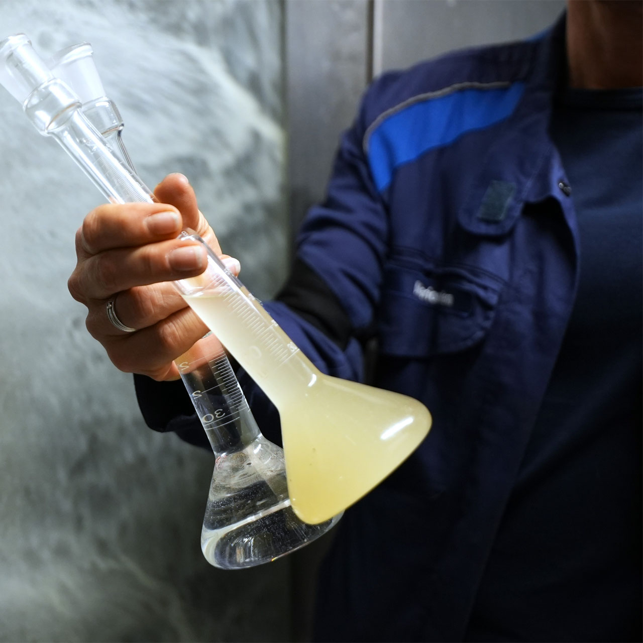 Person hält zwei Glasgefäße mit Flüssigkeiten, eine trüb und gelblich, die andere klar, aus denen Dampf aufsteigt.