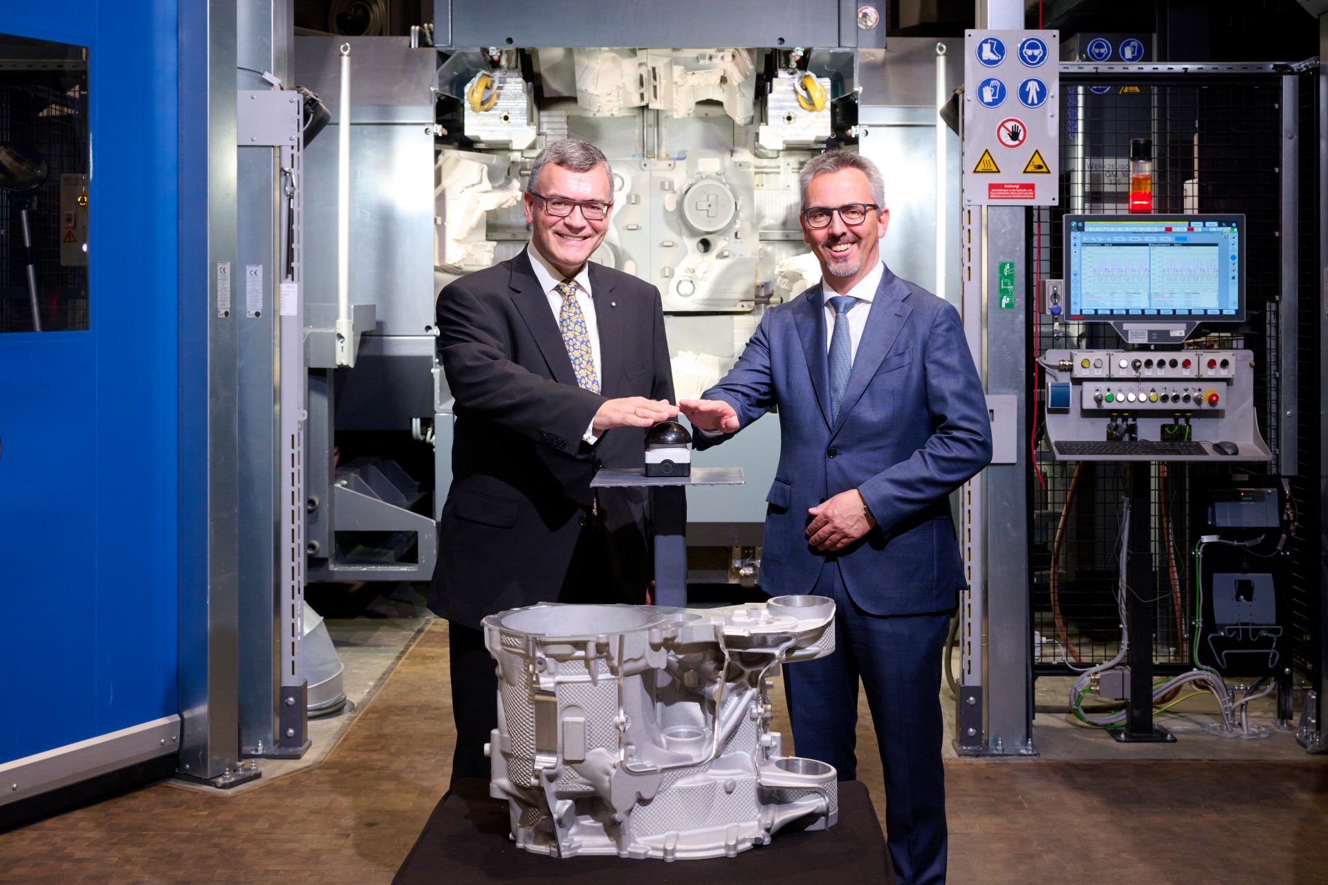 Zwei Männer in Anzügen stehen vor einem neuen E-Motorengehäude und geben sich die Hand 