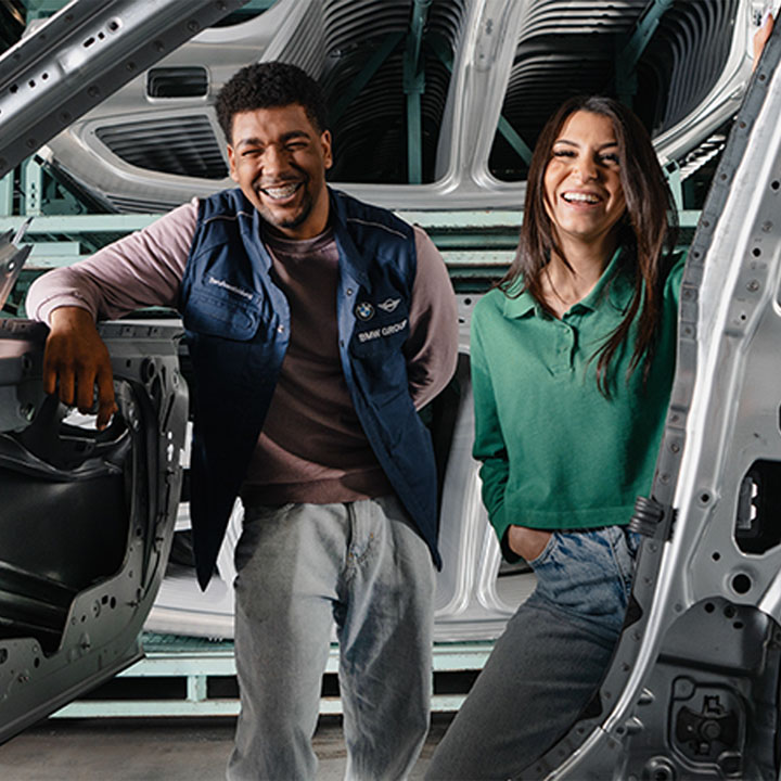 Two student interns stand in production and smile through a vehicle body.