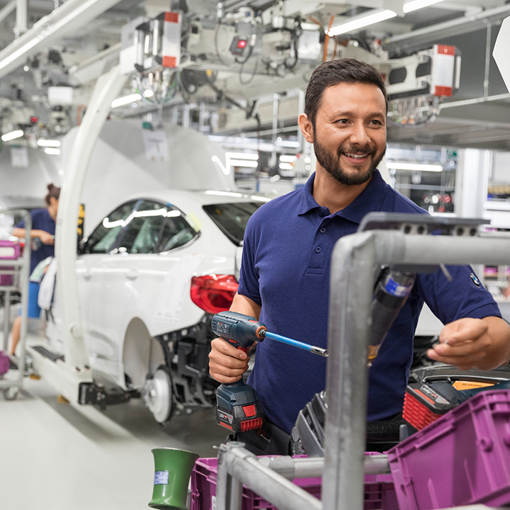 A man works with a screwdriver on vehicle parts with an almost finished car body in the background.