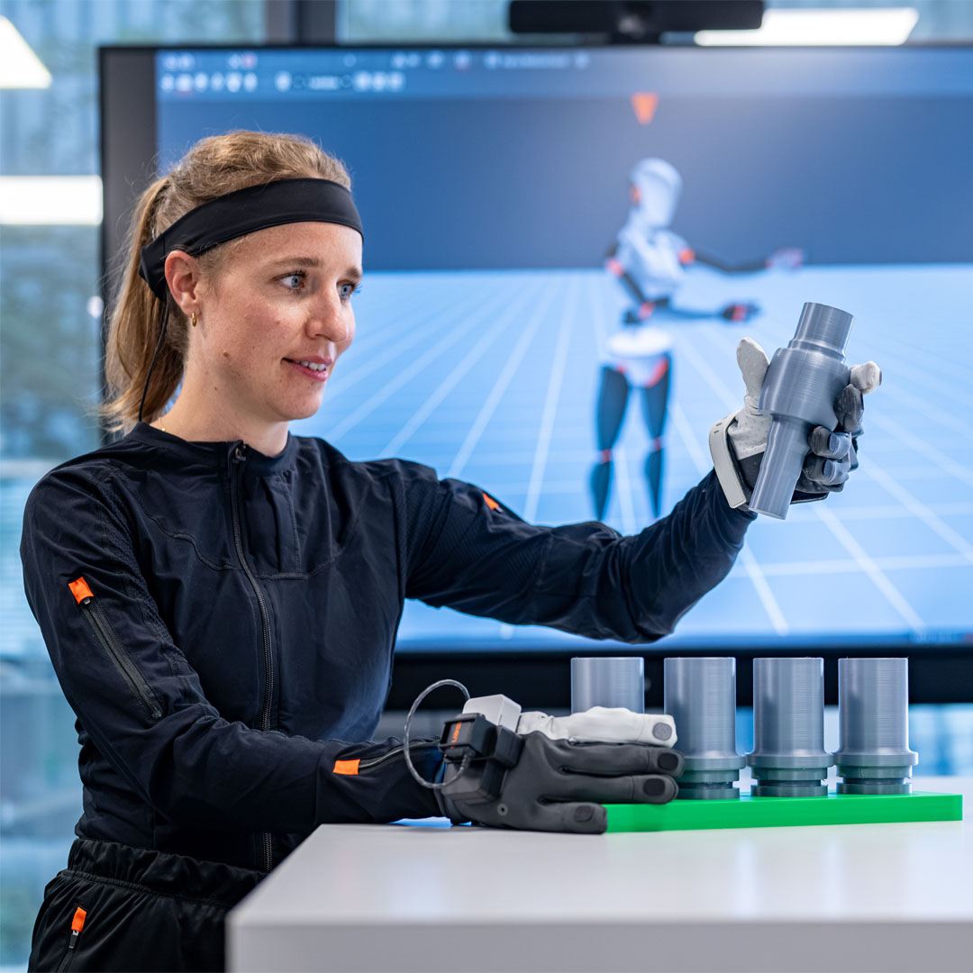 Woman wearing a black headband and black suit with a gray robotic hand holding a gray component in front of a screen showing a humanoid robot.