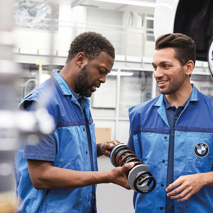 Zwei Mitarbeiter stehen mit einer Feder in der Hand neben einem Auto, welches in die Luft gehoben wurde.
