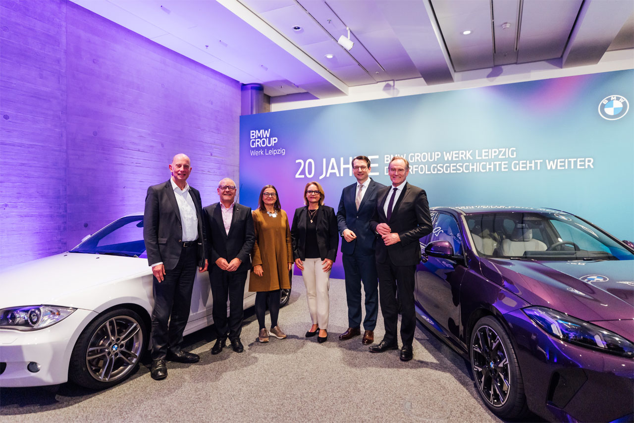 Sechs Personen stehen zwischen einem weißen und einem schwarzen BMW vor einer Wand mit der Aufschrift '20 Jahre BMW Group Werk Leipzig Erfolgsgeschichte geht weiter'.