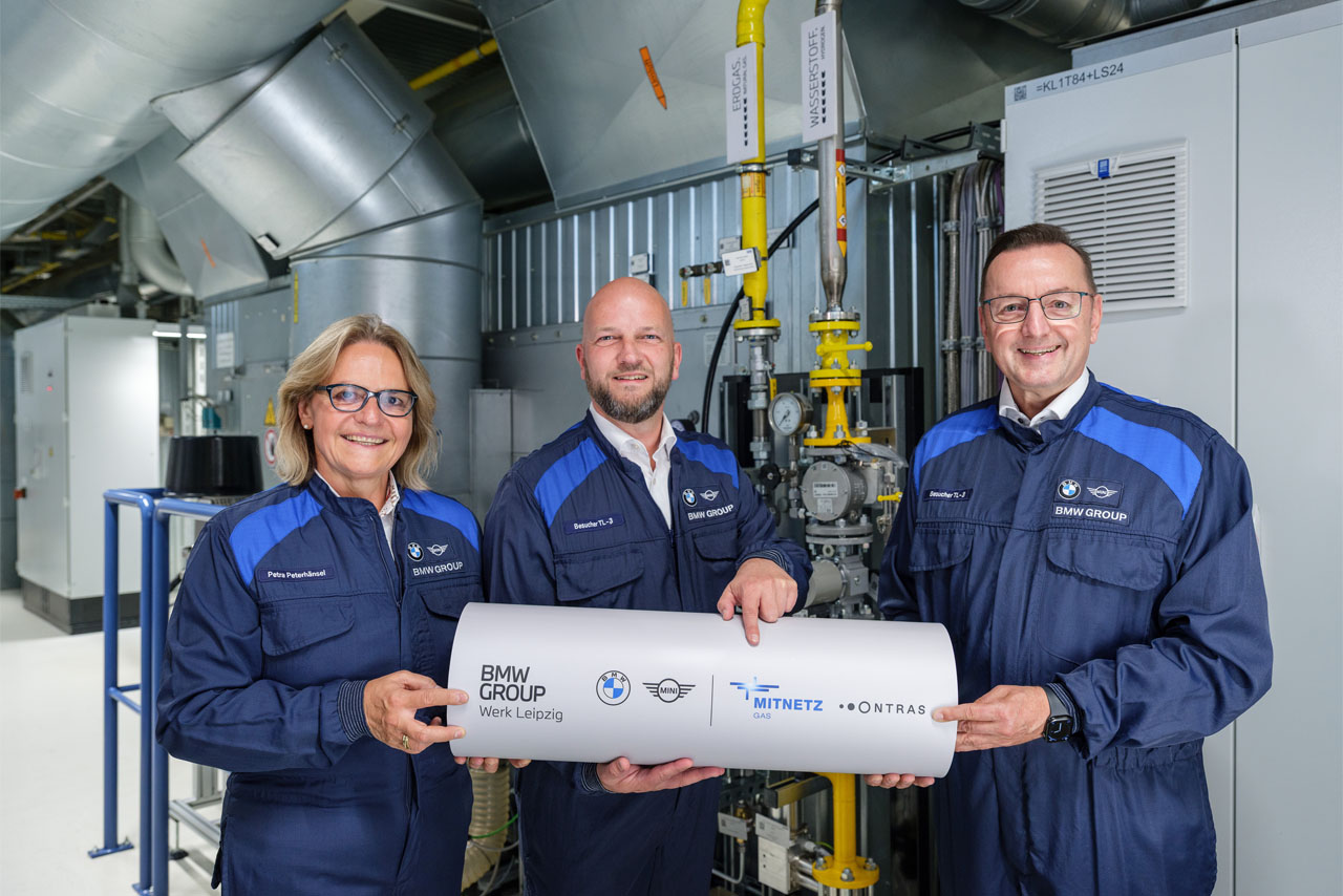 Three people in blue BMW Group work jackets hold a white pipe with BMW Group, Mitnetz, and Ontras logos in a technical facility.