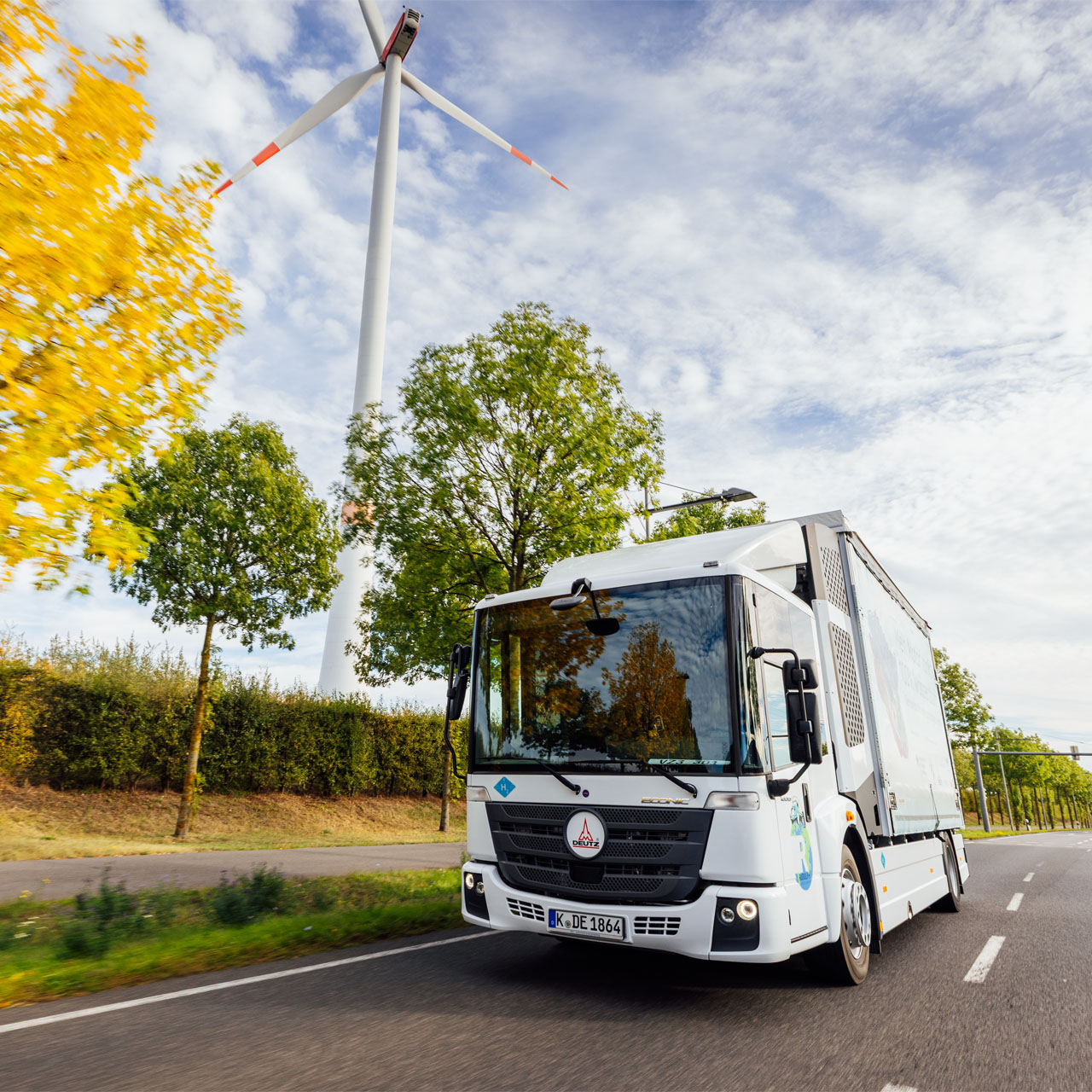 White truck driving on a country road with a wind turbine and trees in the background.