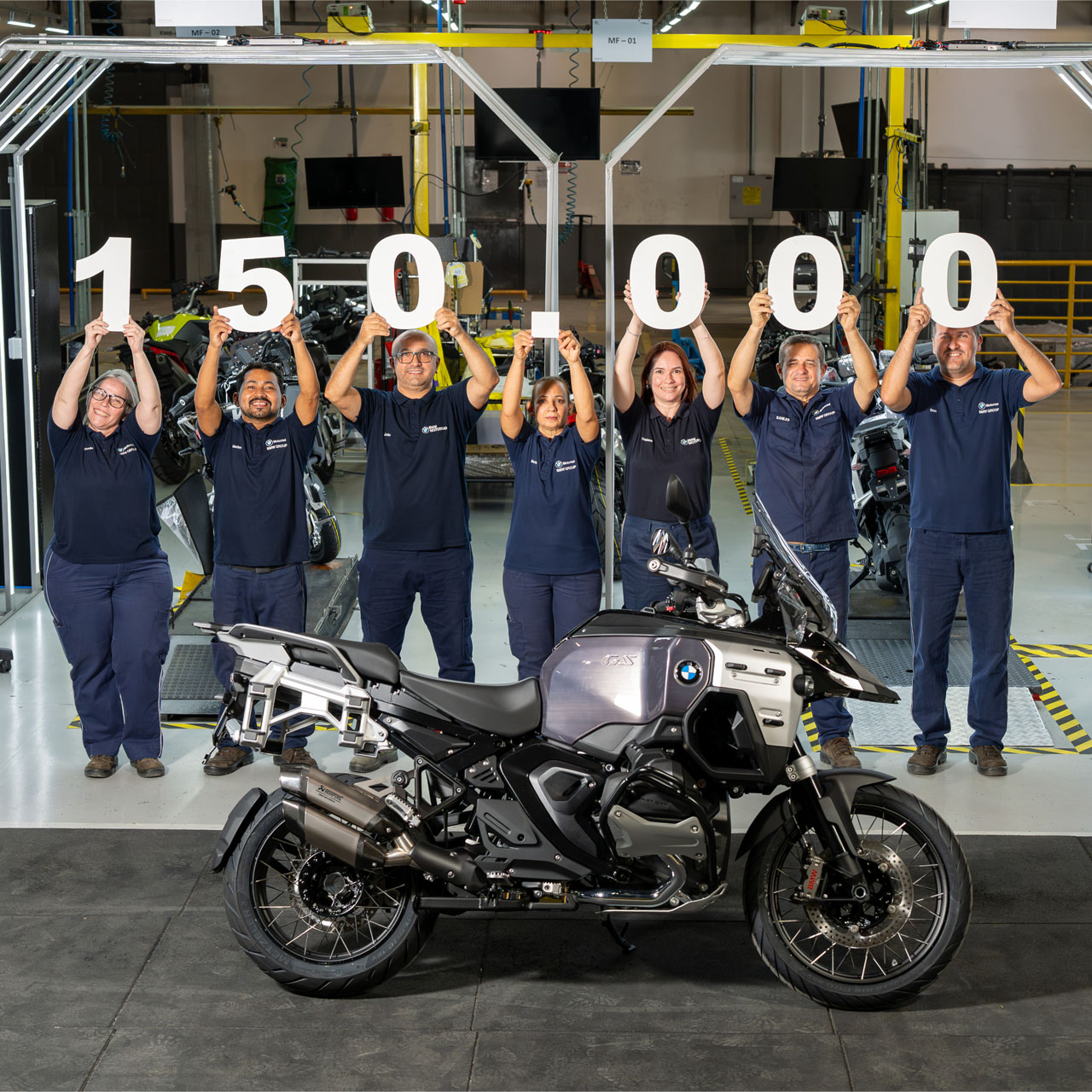 Group of seven people in blue work clothes standing behind a gray BMW motorcycle holding large white numbers forming '150 000'.