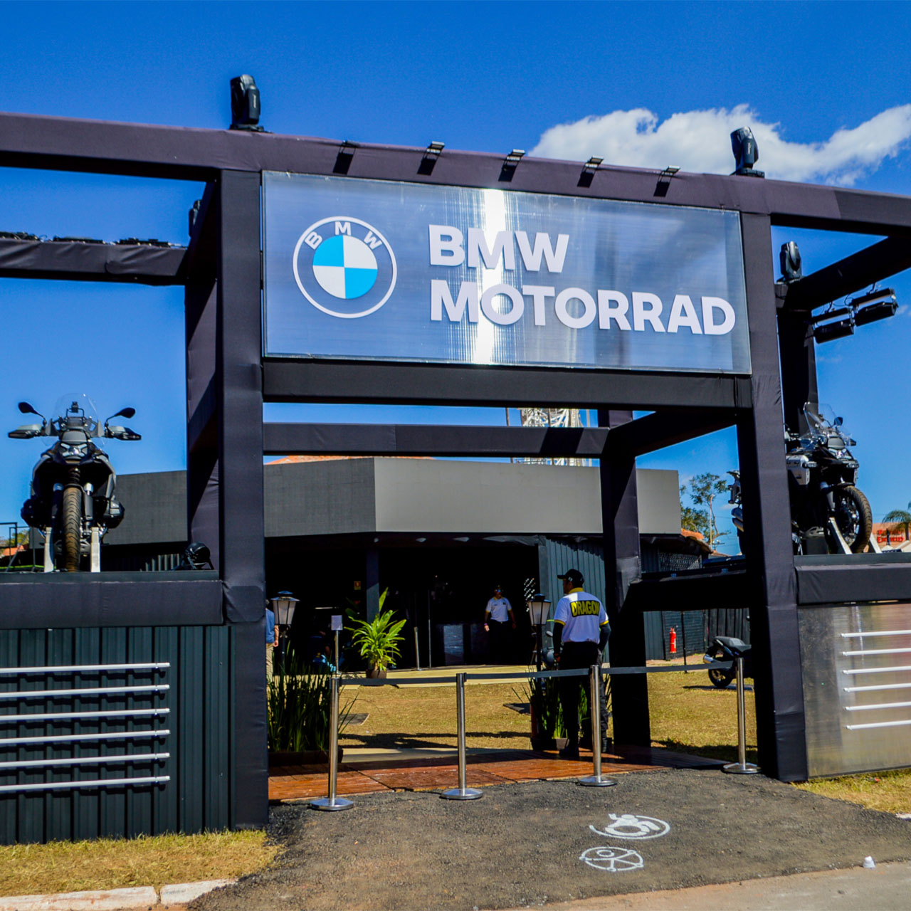 Pavilhão de entrada com o letreiro «BMW MOTORRAD» e duas motos em plataformas, sob um céu ensolarado.