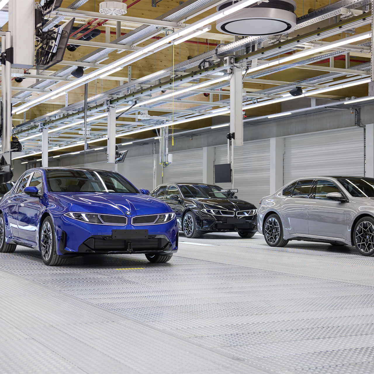 Zwei BMW-Elektroautos in Blau und Silber stehen in einer modernen Produktionshalle mit hellen Deckenleuchten und sichtbaren Rohrleitungen.