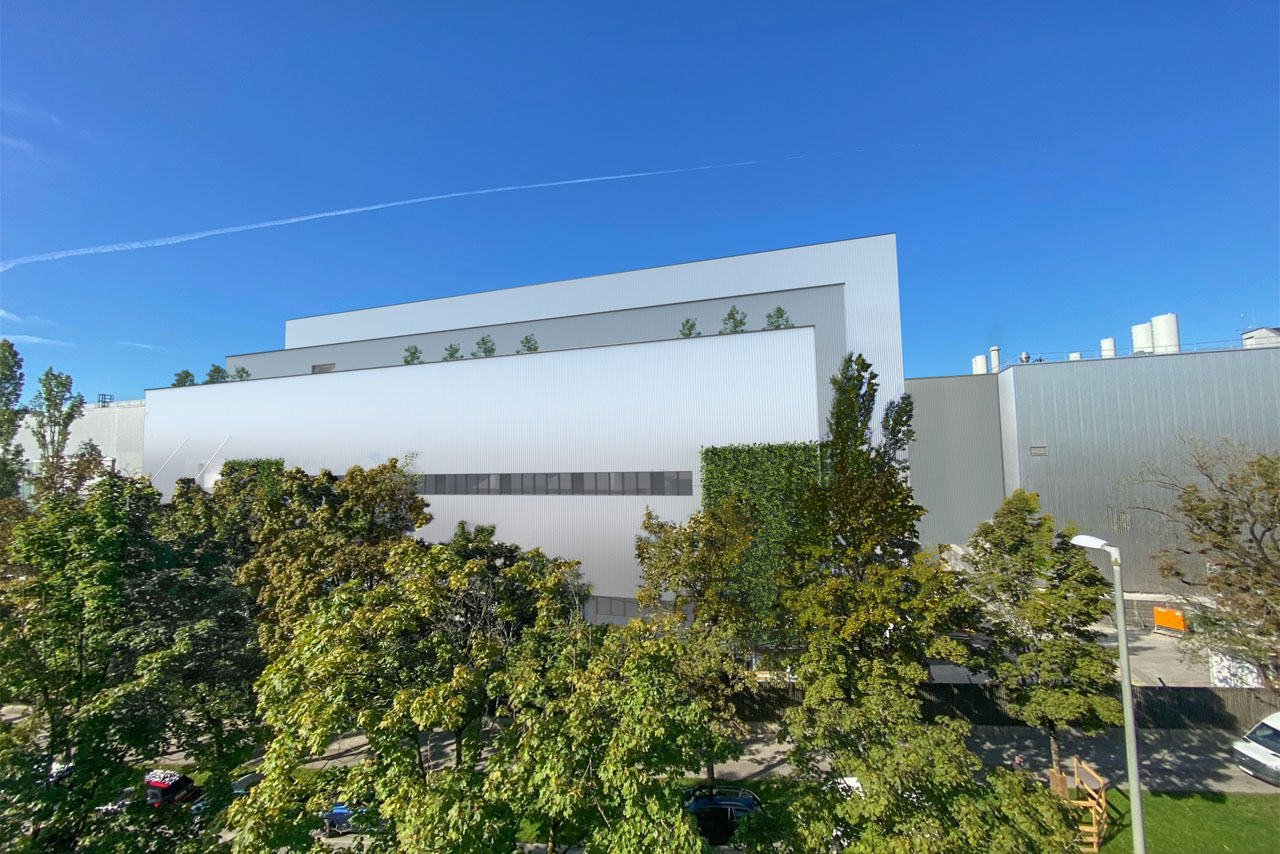 Großes, modernes weißes Gebäude mit rechteckigen Fenstern, umgeben von grünen Bäumen unter blauem Himmel.