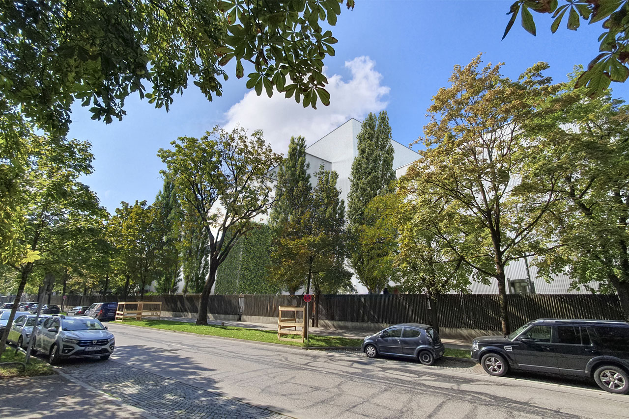 Straße mit parkenden Autos und Bäumen vor einem weißen, mehrstöckigen Gebäude unter blauem Himmel mit wenigen Wolken.