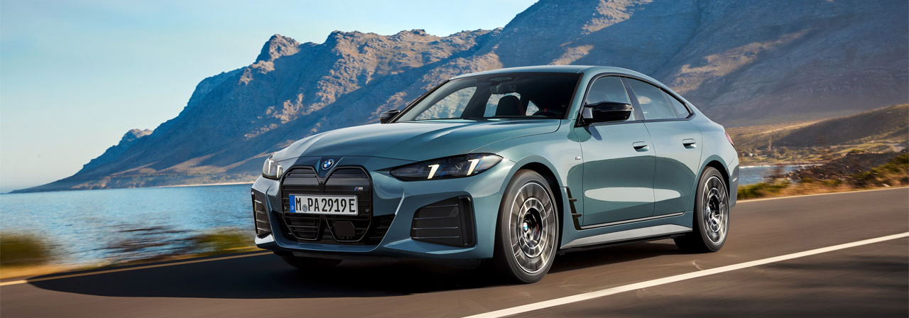 Grauer BMW i4 fährt auf einer Landstraße am Meer entlang mit Bergen im Hintergrund.