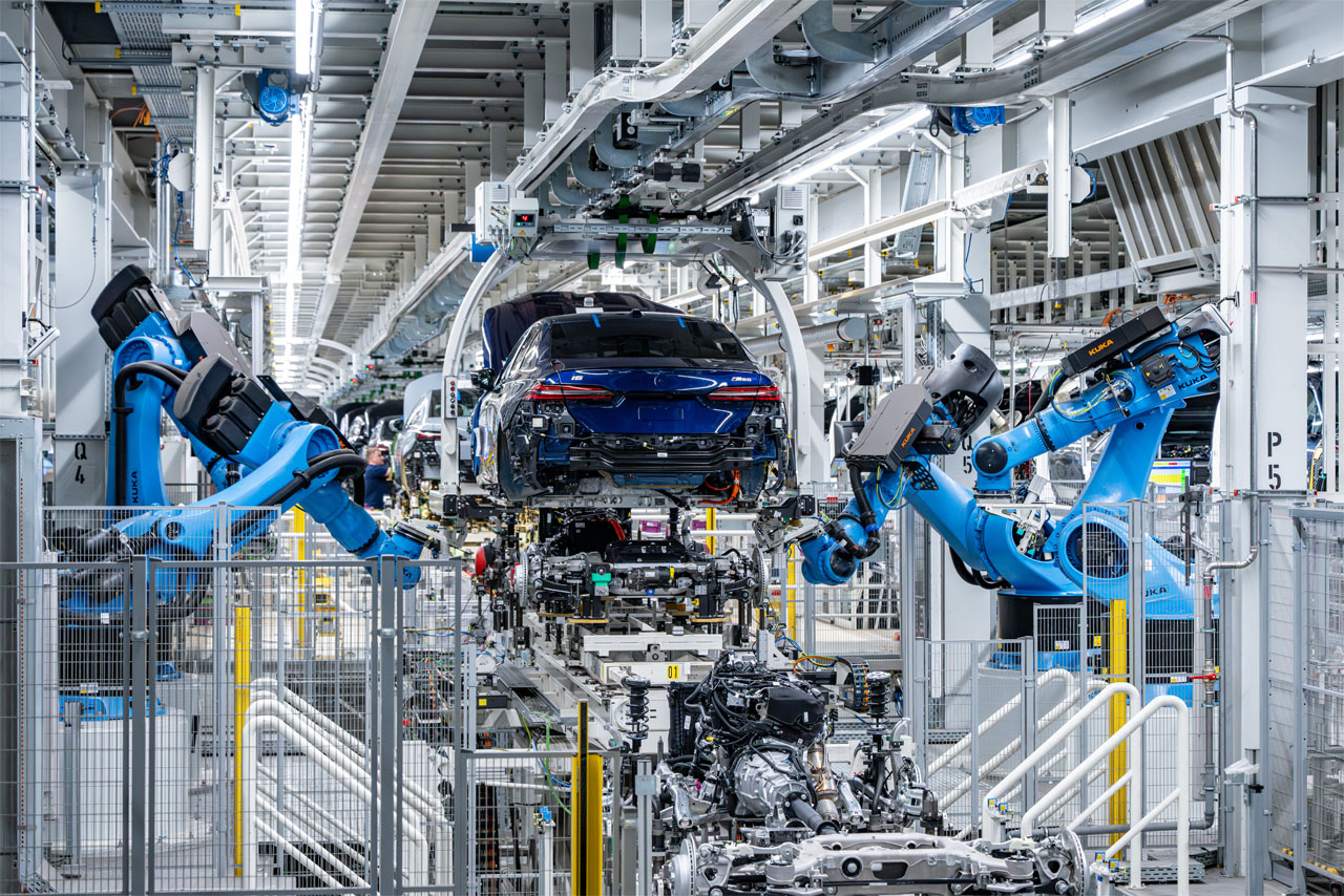 Fabrikhalle mit zwei großen Industrierobotern, die an einem blauen Autoheck arbeiten.