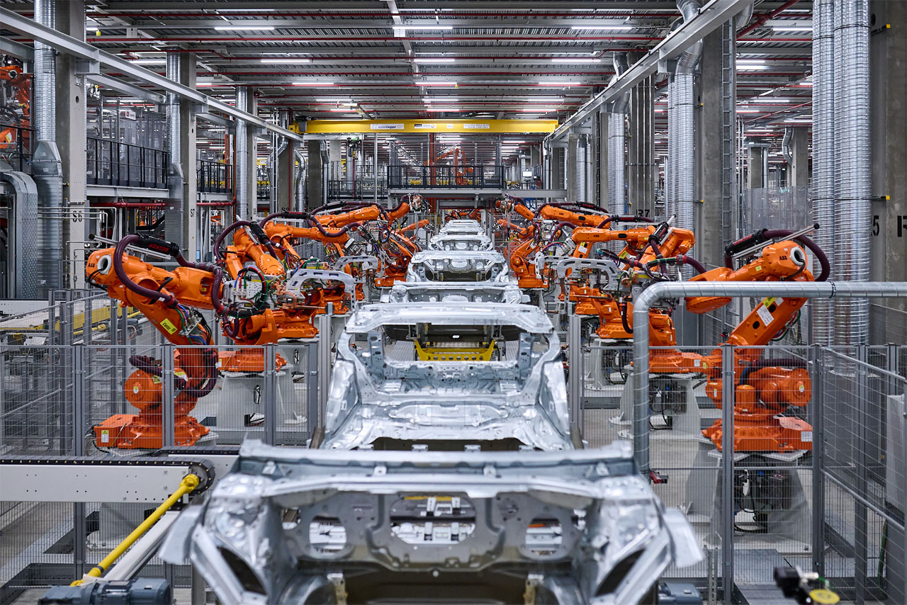 A manufacturing facility with several orange industrial robots working on a line of vehicle bodies.