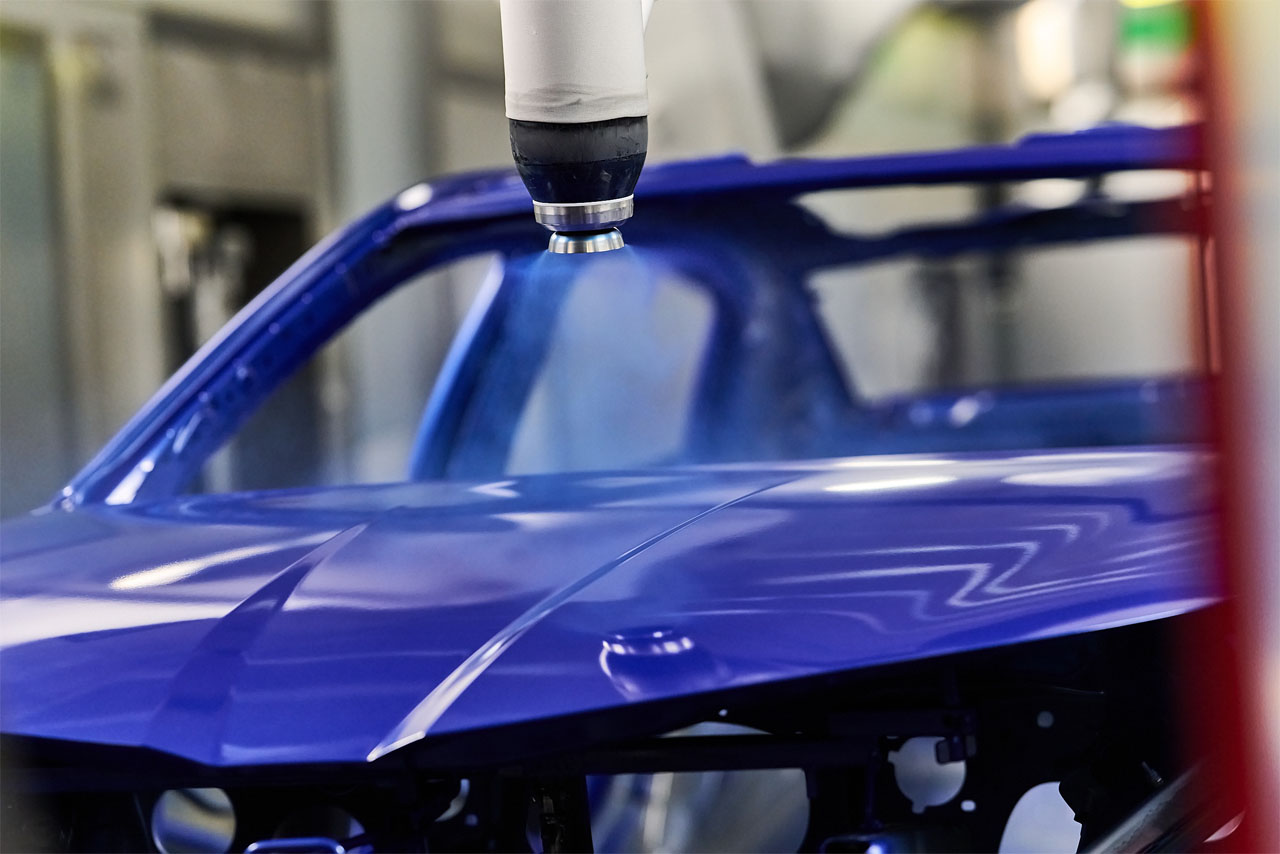 A close-up of a blue car roof in a paint shop, with a spray gun hovering above it.