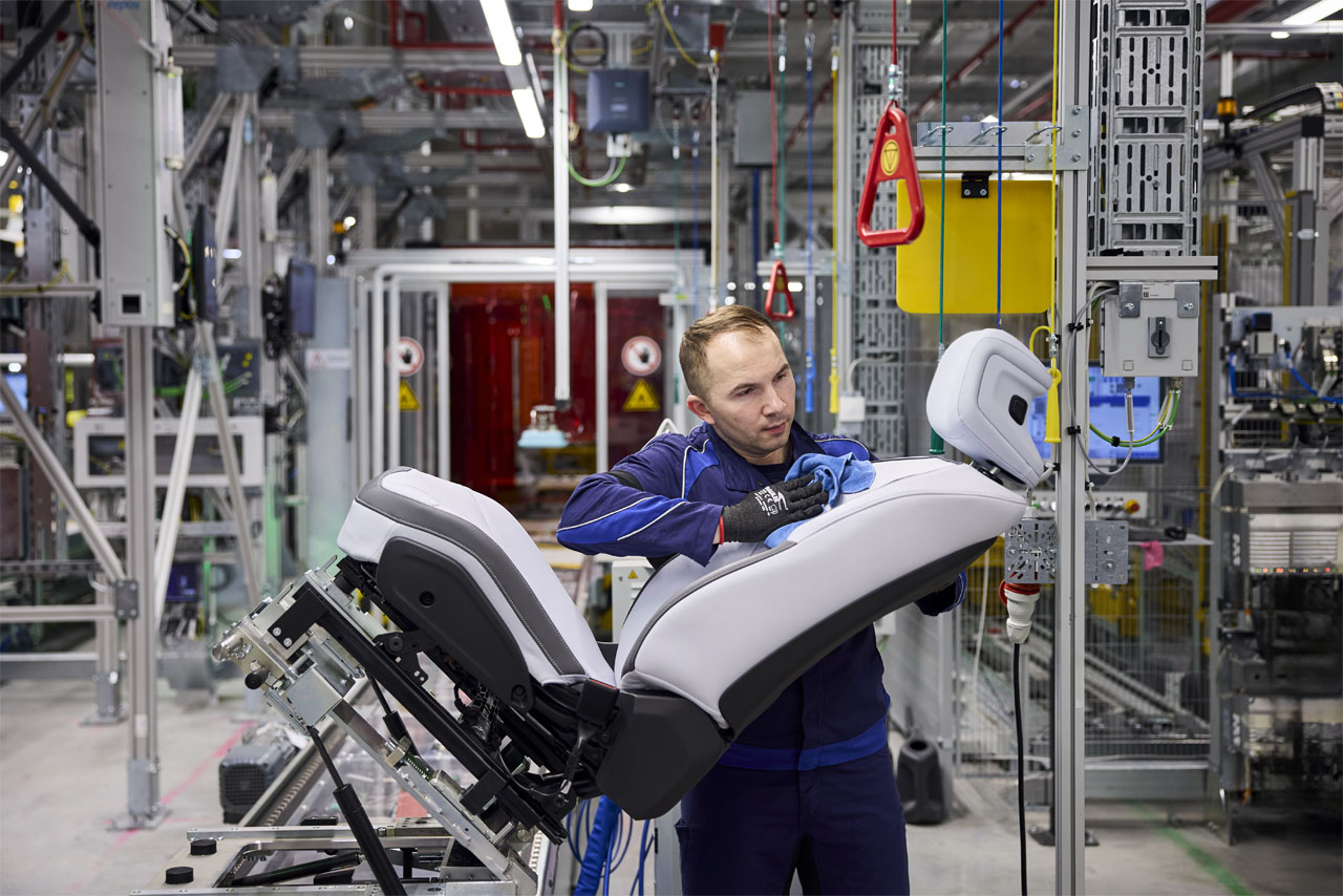 A man in work clothes is installing a car seat assembly in a factory with production equipment in the background.