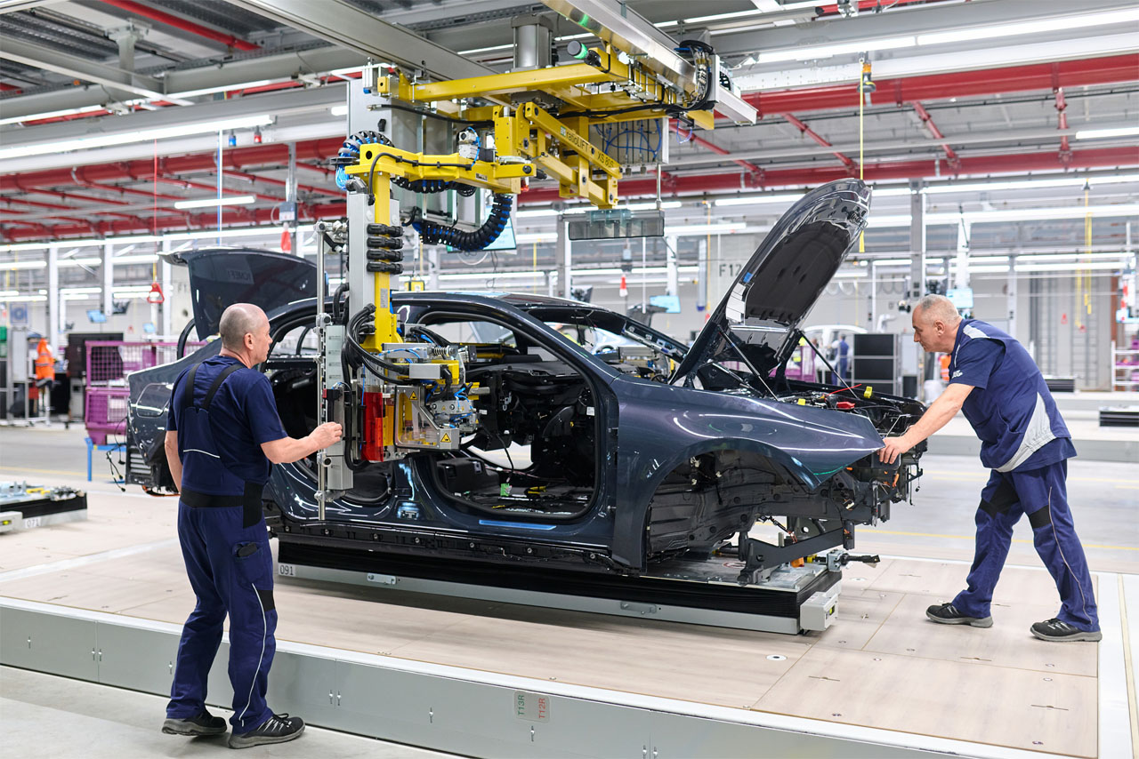 In a factory, two men are assembling a black car body with the hood open on an assembly line.