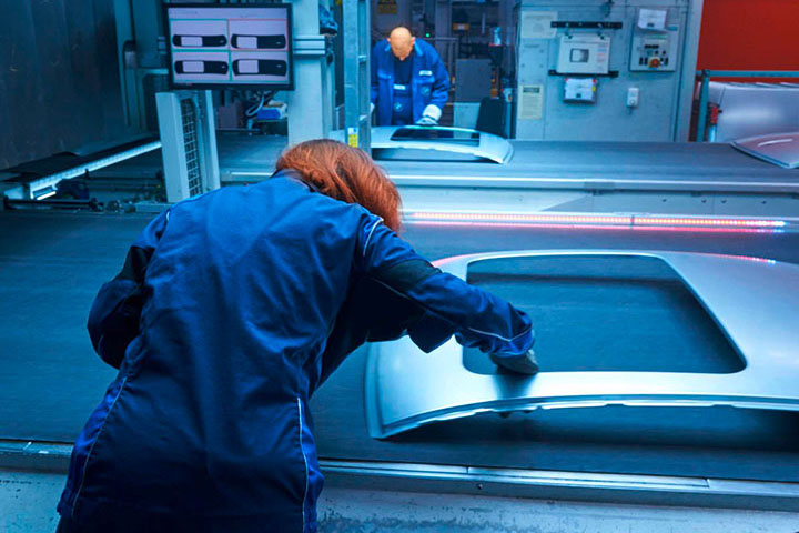 A woman with red hair and a blue bodysuit working on a car part in the press shop.
