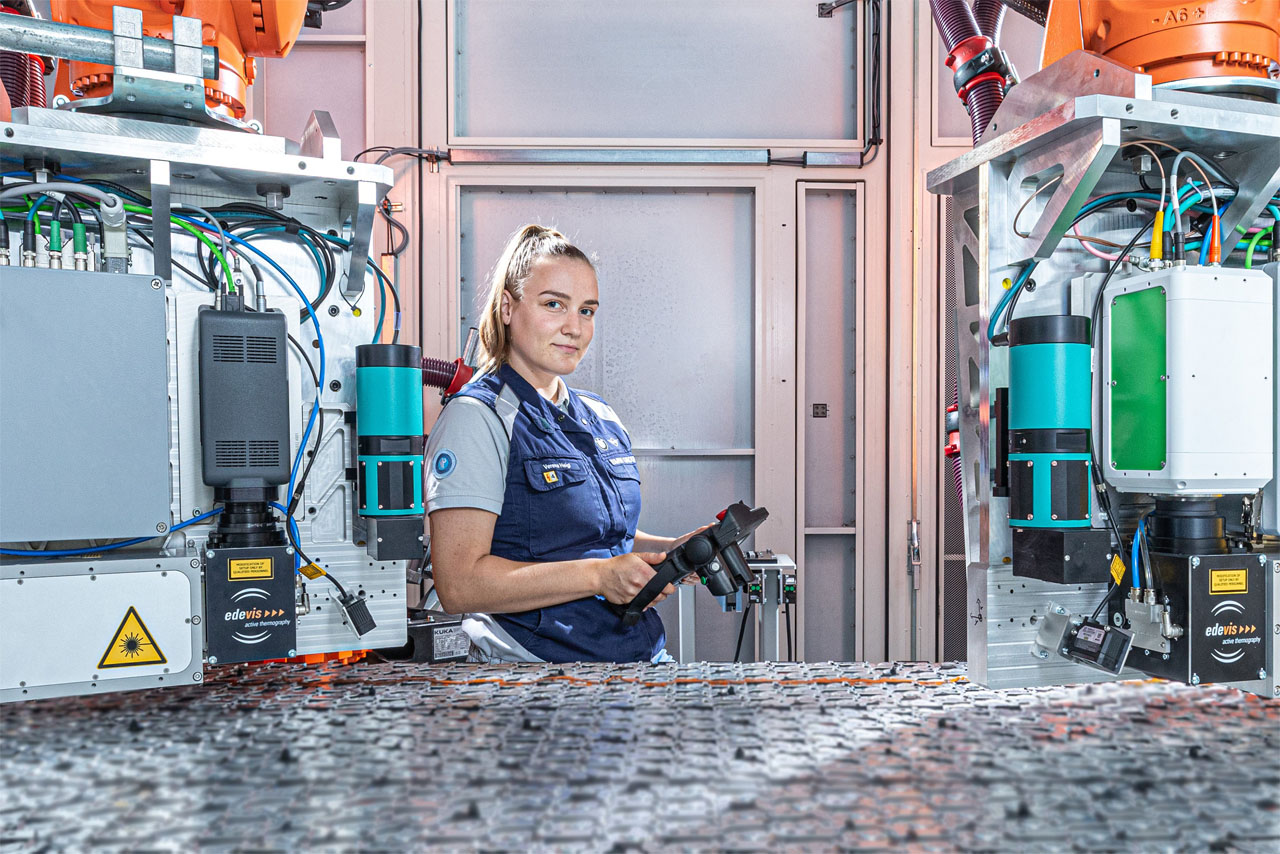 A woman with blonde hair and a control unit stands in the middle of a battery cell room, surrounded by several machines.