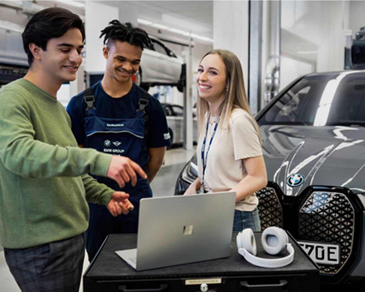 Drei Schüler*innen stehen zusammen in einer Werkstatt vor einem BMW iX und besprechen etwas am Computer.
